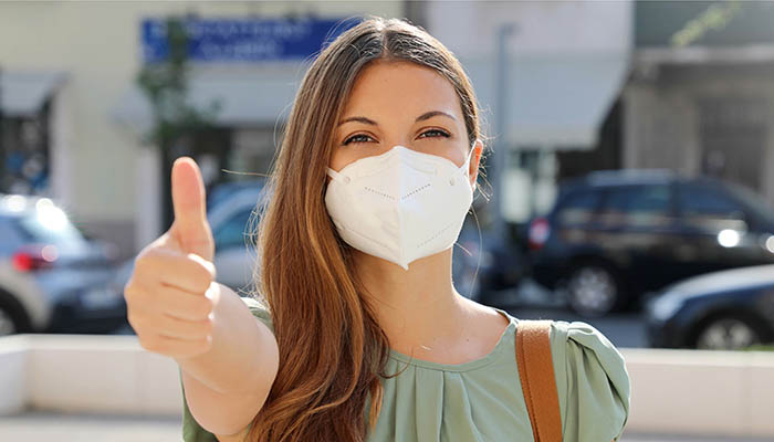 COVID-19 Positive young woman wearing protective mask KN95 FFP2 avoiding Coronavirus disease 2019 showing thumbs up in city street