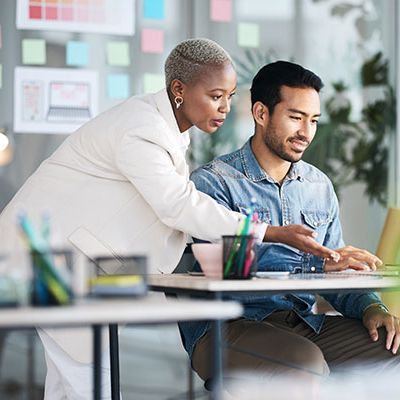 In einem professionellen Setting diskutieren ein Mann und eine Frau über Mitarbeiterfeedback. Der Mann sitzt am Tisch und zeigt seiner Kollegin Führungskräftefeedbacks von seinem Laptop aus.