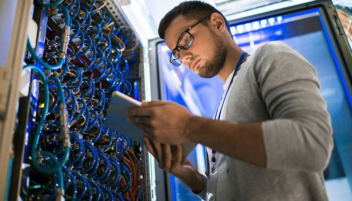 Low angle  portrait of young man using digital tablet standing by server cabinet while working with supercomputer in blue light