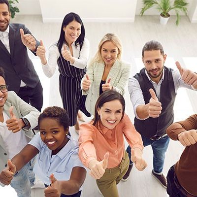 Eine Gruppe glücklicher Mitabeitenden lächelt in die Kamera und zeigt dabei durch Daumen nach oben ihre Mitarbeiterzufriedenheit.