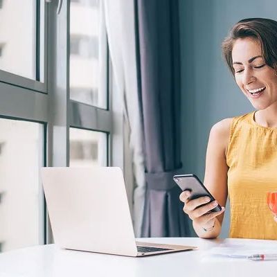 Dame schaut auf das Handy mit Caffè in der Hand, gegenüber einen Rechner