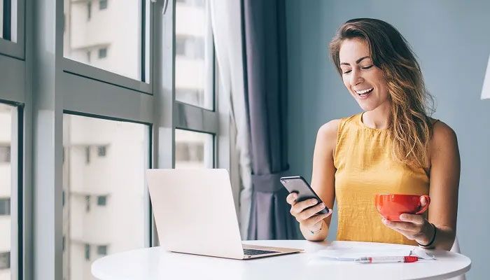 Dame schaut auf das Handy mit Caffè in der Hand, gegenüber einen Rechner