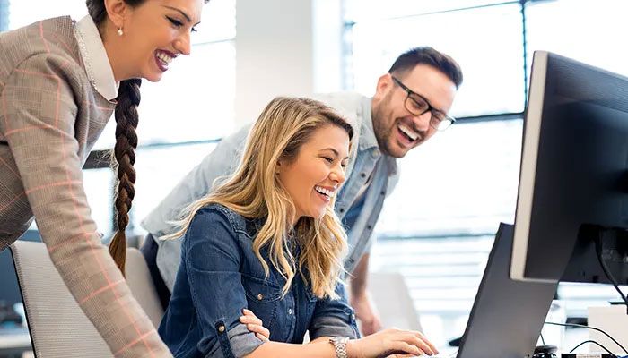 Drei Mitarbeiter und Mitarbeiterinnen schauen lächelnd auf einen Laptop und bewerten die Mitarbeiterbefragung in Behörden.