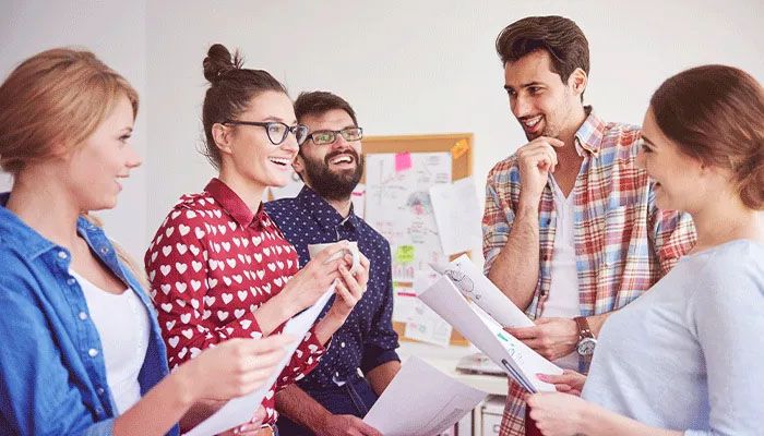 Team-Monitor: Fünf Personen stehen im Kreis mit Blättern in der Hand und lachen.