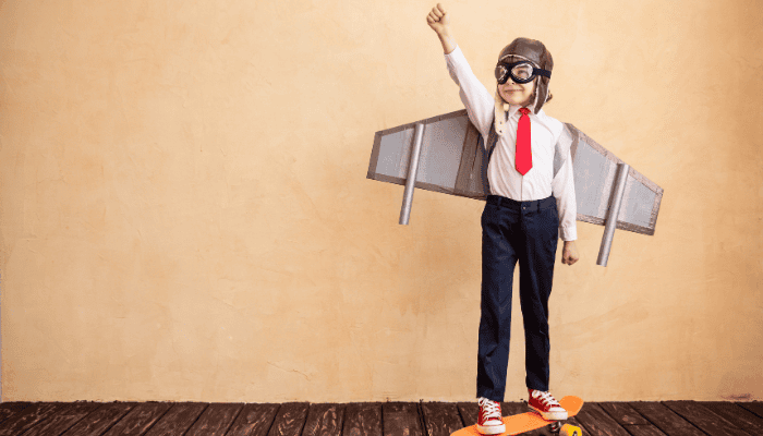 Ein kleiner Junge in Superman Pose und angeklebten Pappflügeln tut lächelnd so, als ob er fliegen würde.