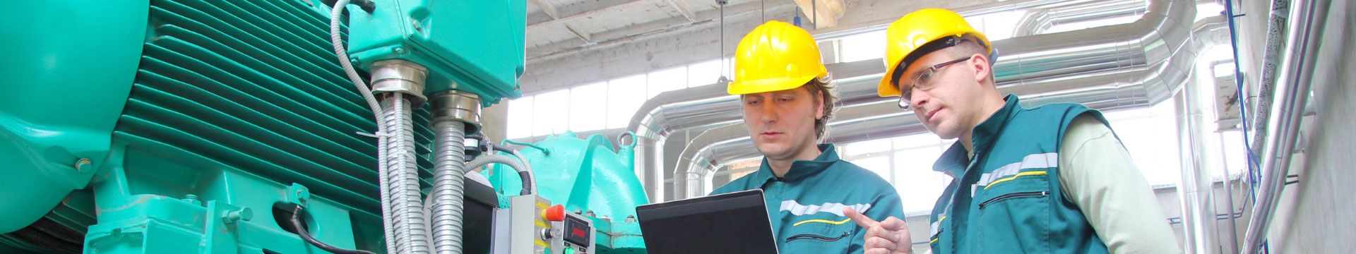 Das Foto zeigt zwei Menschen mit einem gelben Helm auf dem Kopf, welche vor Industriemaschinen stehen