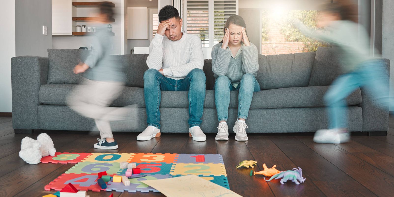 Shot of two young parents looking tired while their children run around at home.