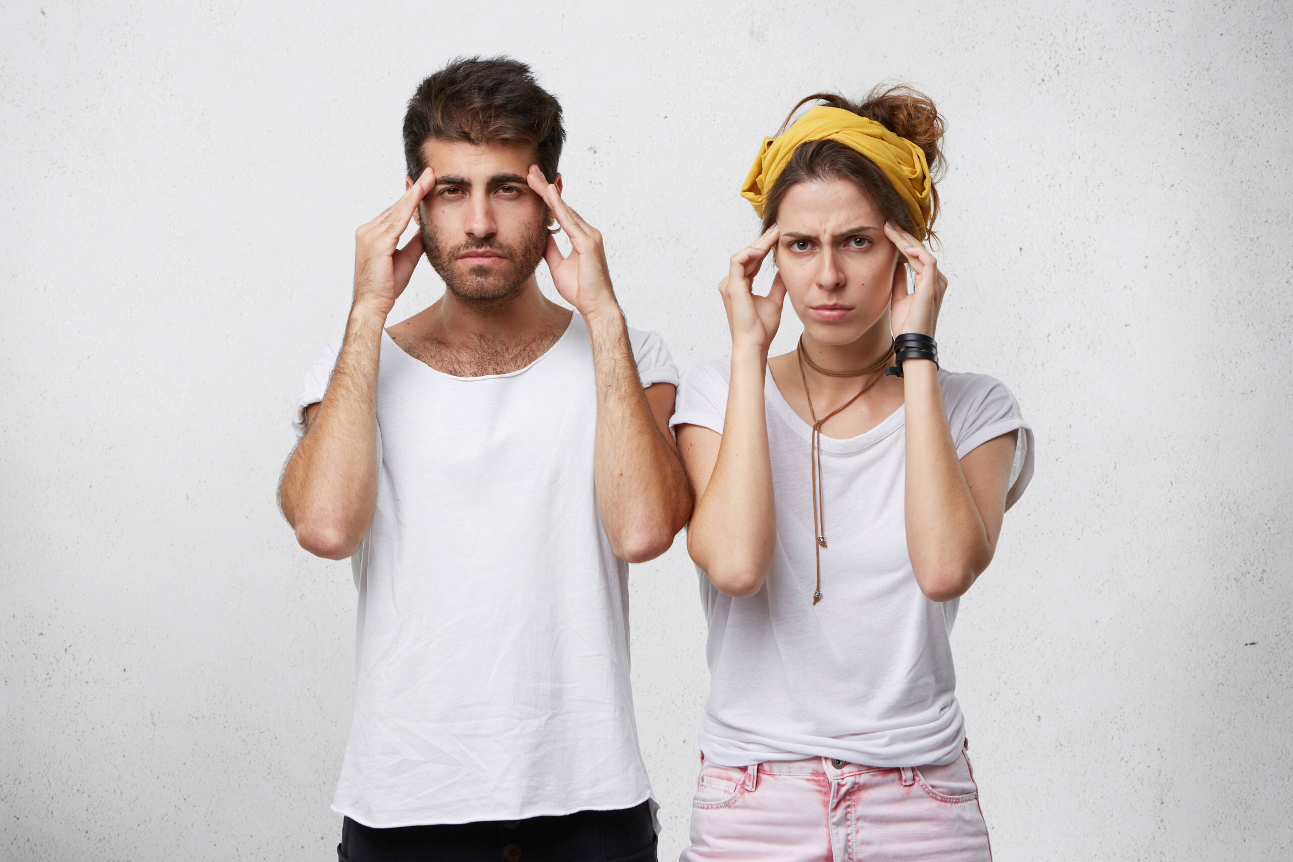 Portrait of bearded handsome man and beautiful woman with yellow headband on head trying to concentrate holding thier hands on temples. Female and male in white clothes remembering something important