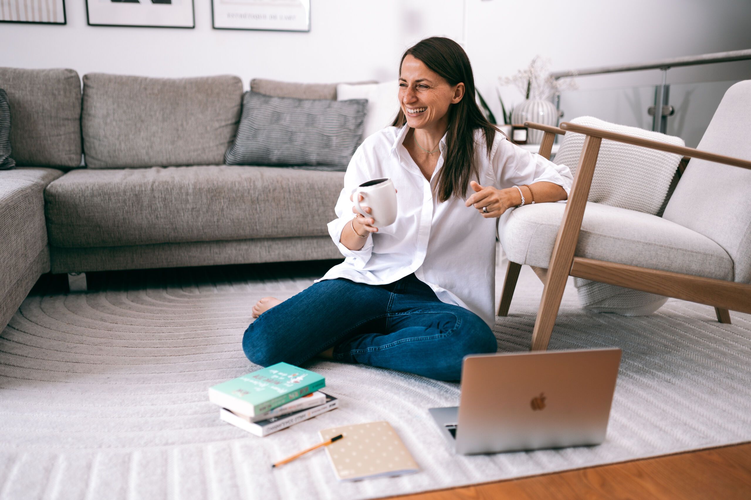 Magdalena Ochs lachend, auf dem Teppichboden sitzend, mit einer Tasse in der Hand, Bücher liegen um die herum, ein Laptop aufgeklappt neben ihr