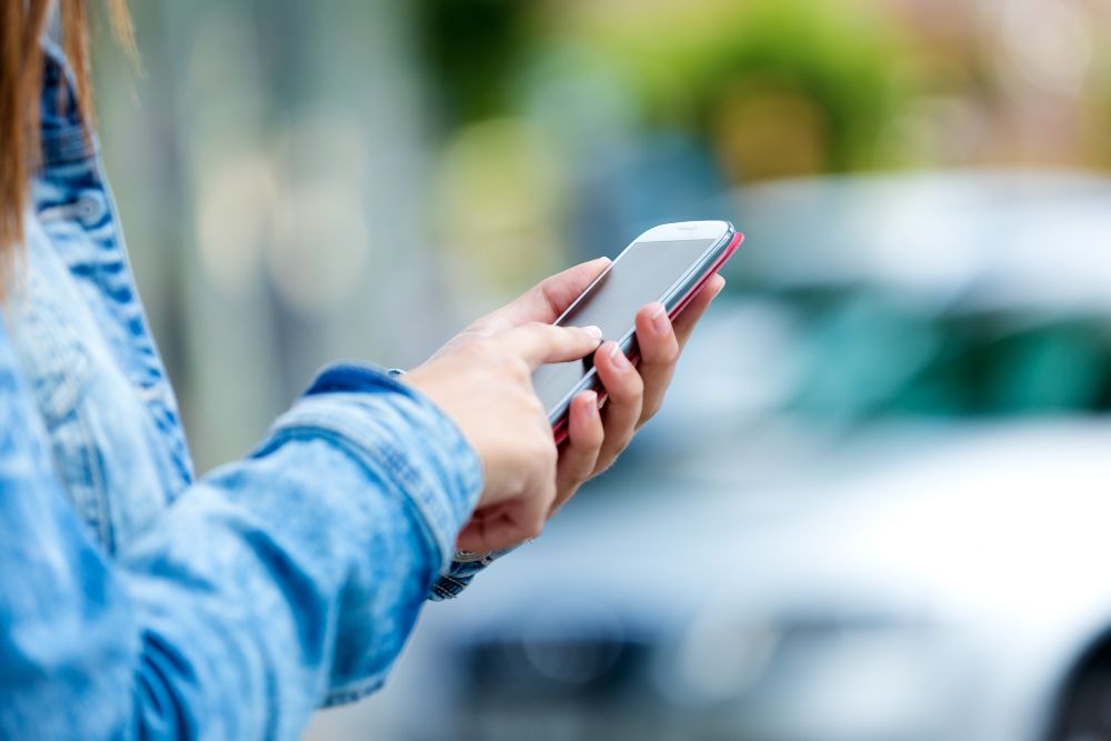 Handy in der Hand einer Frau vor parkenden Autos.