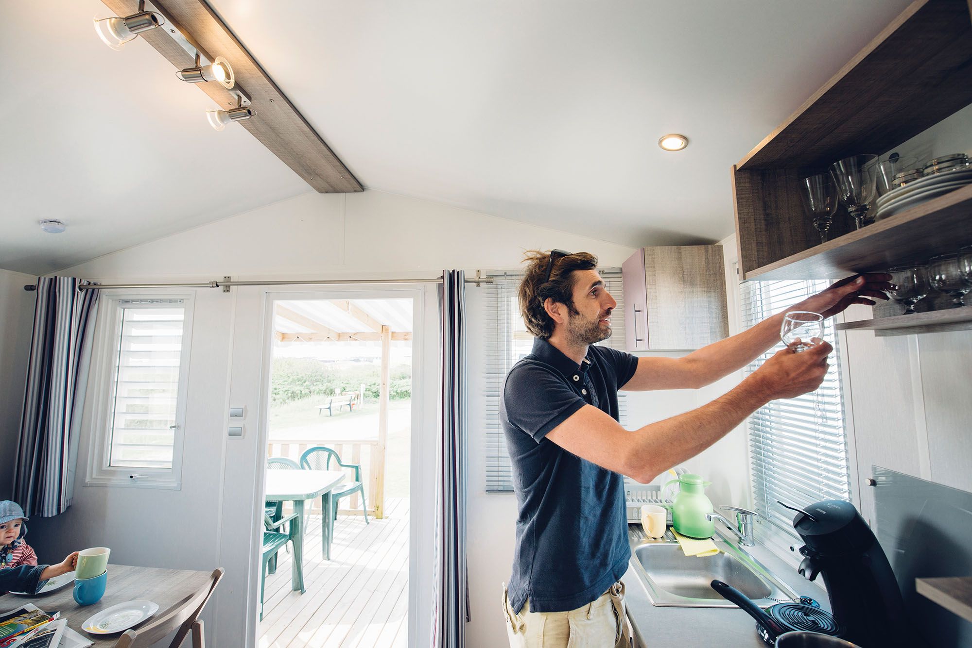 Mann holt in der Küche eines kleinen Mobilheim auf dem Campingplatz Rantum ein Glas aus dem Schrank