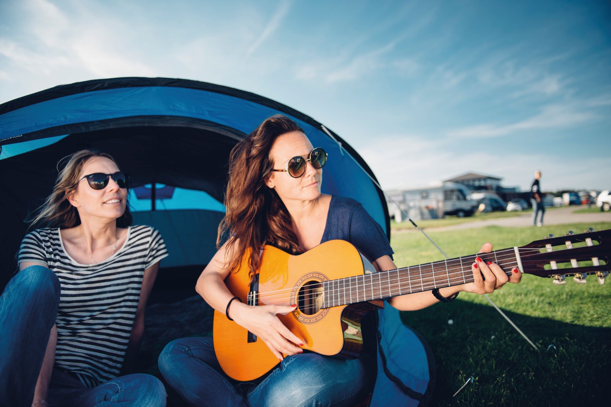 Frau spielt Gitarre und sitzt dabei mit einer anderen Frau im Eingang zu ihrem Zelt