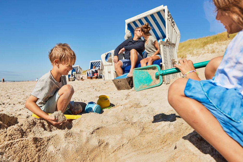Frau und Mann küssen sich im Strandkorb während Junge und Mädchen davor im Sand spielen