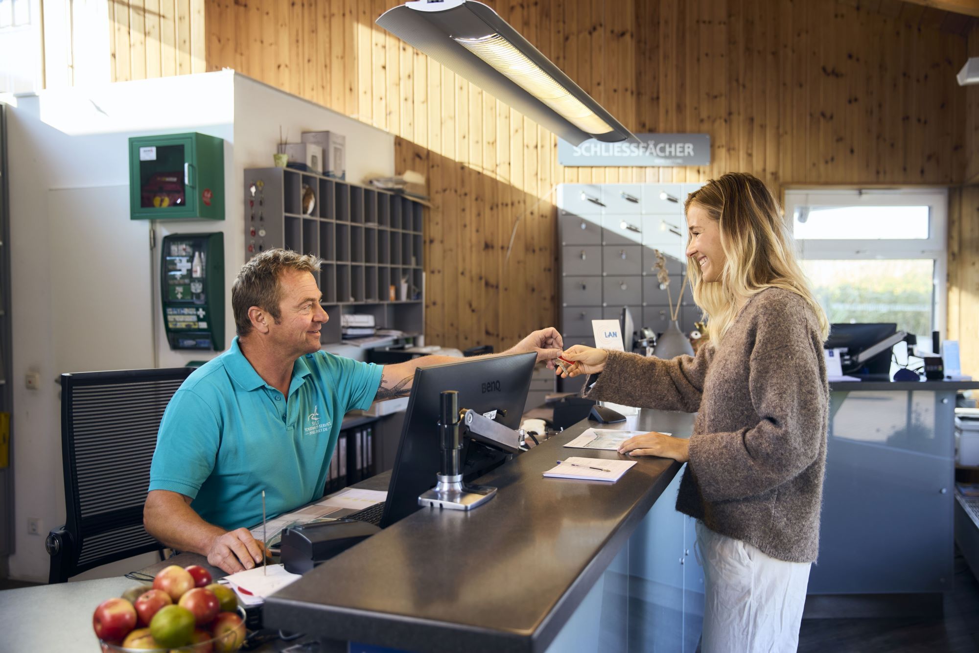 Männlicher Mitarbeiter in türkisfarbenem Poloshirt des Campingplatzes reicht einem weiblichen Gast die Schlüssel zu ihrer Unterkunft