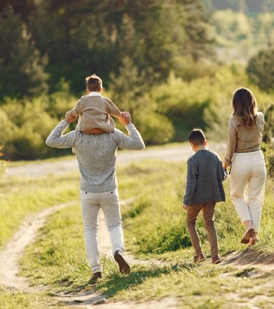 Familie beim Spaziergang an einem sonnigen Herbsttag – Mutter hält die Hand des älteren Sohnes, Vater trägt den kleinen Sohn auf den Schultern, umgeben von herbstlicher Natur.