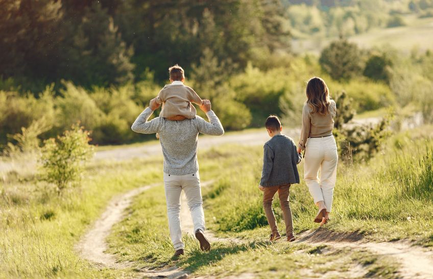 Familie beim Spaziergang an einem sonnigen Herbsttag – Mutter hält die Hand des älteren Sohnes, Vater trägt den kleinen Sohn auf den Schultern, umgeben von herbstlicher Natur.