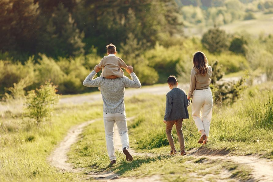 Familie beim Spaziergang an einem sonnigen Herbsttag – Mutter hält die Hand des älteren Sohnes, Vater trägt den kleinen Sohn auf den Schultern, umgeben von herbstlicher Natur.