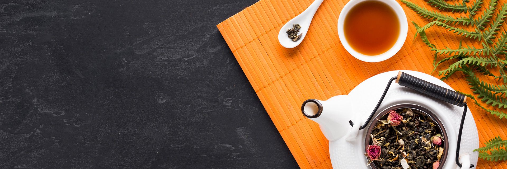 japanische Teezeremonie. Auf einer Bambusmatte steht eine offene Teekanne mit Teeblättern im Sieb, dazu eine Teetasse mit bernsteinfarbenem Tee und ein Löffel mit ein paar losen Teeblättern. Daneben ein grüner Zweig.