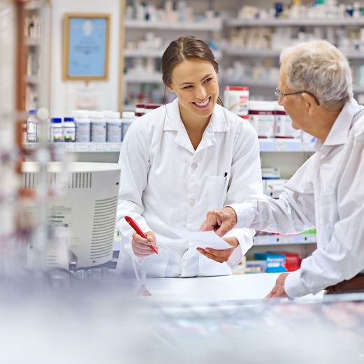 zwei Personen stehen in einer Apotheke. Die Person hinter dem Tisch zeigt der Person vor dem Tisch etwas auf einem Papier.