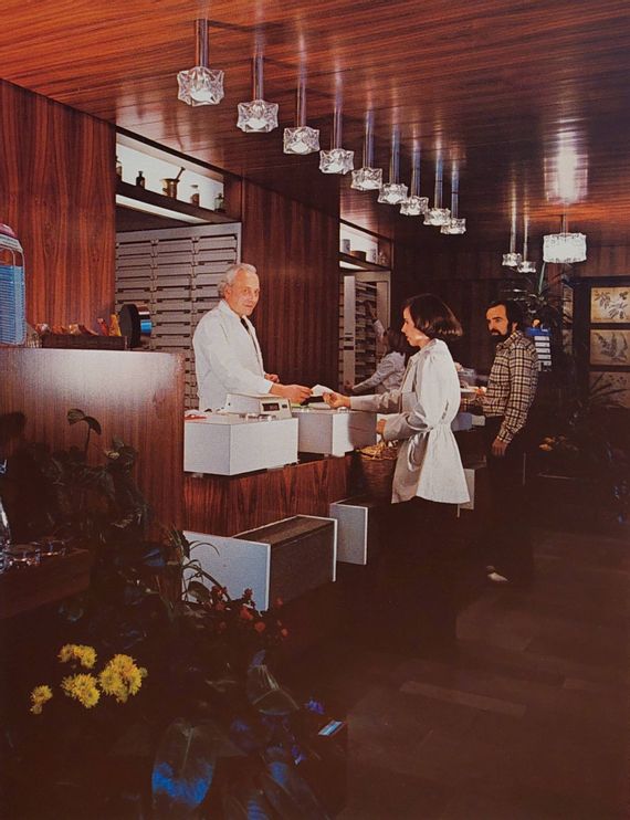 Innenansicht der Schlossapotheke in den 80er Jahren. Zwei Kunden stehen in der Apotheke, der Apothekenleiter händigt gerade eine Quittung aus.