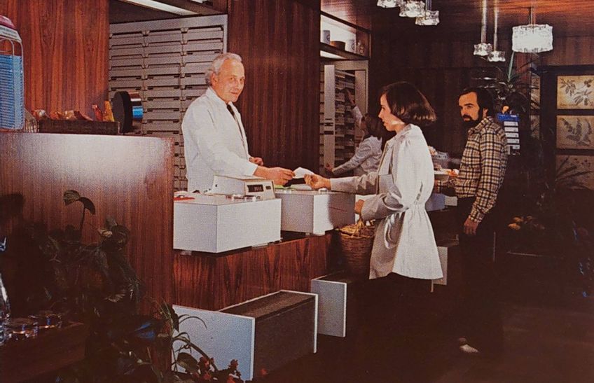 Innenansicht der Schlossapotheke in den 80er Jahren. Zwei Kunden stehen in der Apotheke, der Apothekenleiter händigt gerade eine Quittung aus.