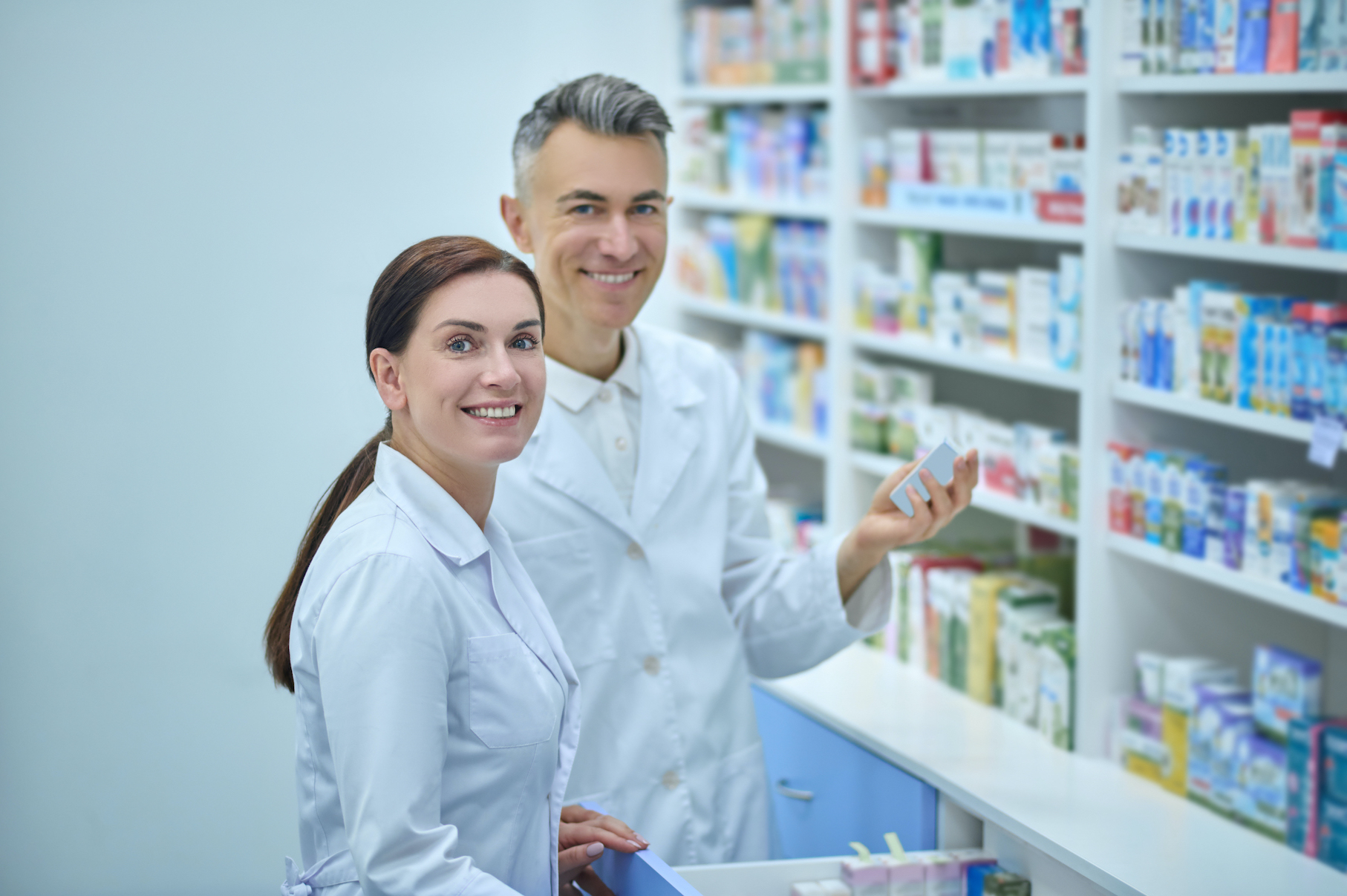 zwei Personen stehen an einem Arzneimittelregal vor einer geöffeten Schublade, die rechte Person hat eine Packung in der Hand