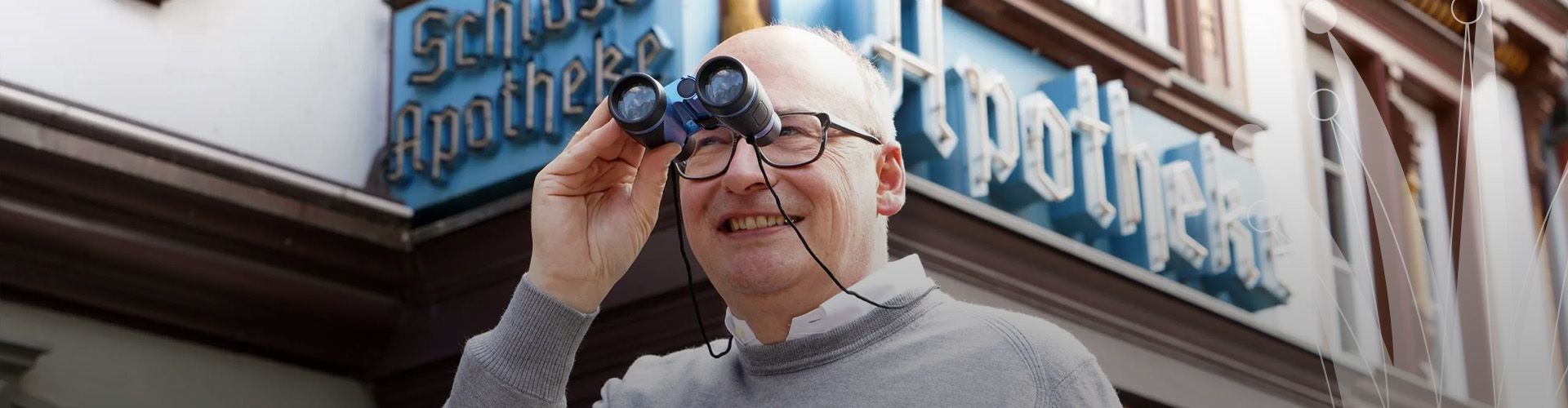 Dr. Gubitz schaut in ein Fernglas, im Hintergrund die Leuchtreklame an der Fassade der Schlossapotheke