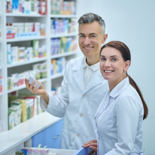 zwei Personen stehen an einem Arzneimittelregal vor einer geöffeten Schublade, die linke Person hat eine Packung in der Hand