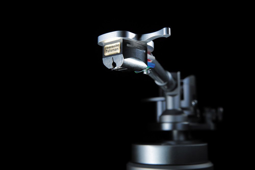Nahaufnahme des clearaudio „Talisman“ Tonabnehmers, der an einem silbernen Tonarm montiert ist. Die Szene ist vor einem tiefschwarzen Hintergrund fotografiert. Das goldene Frontplättchen des Tonabnehmers setzt einen glänzenden Akzent, und die farbigen Anschlusskabel am hinteren Ende des Systems sind minimal zu erkennen.