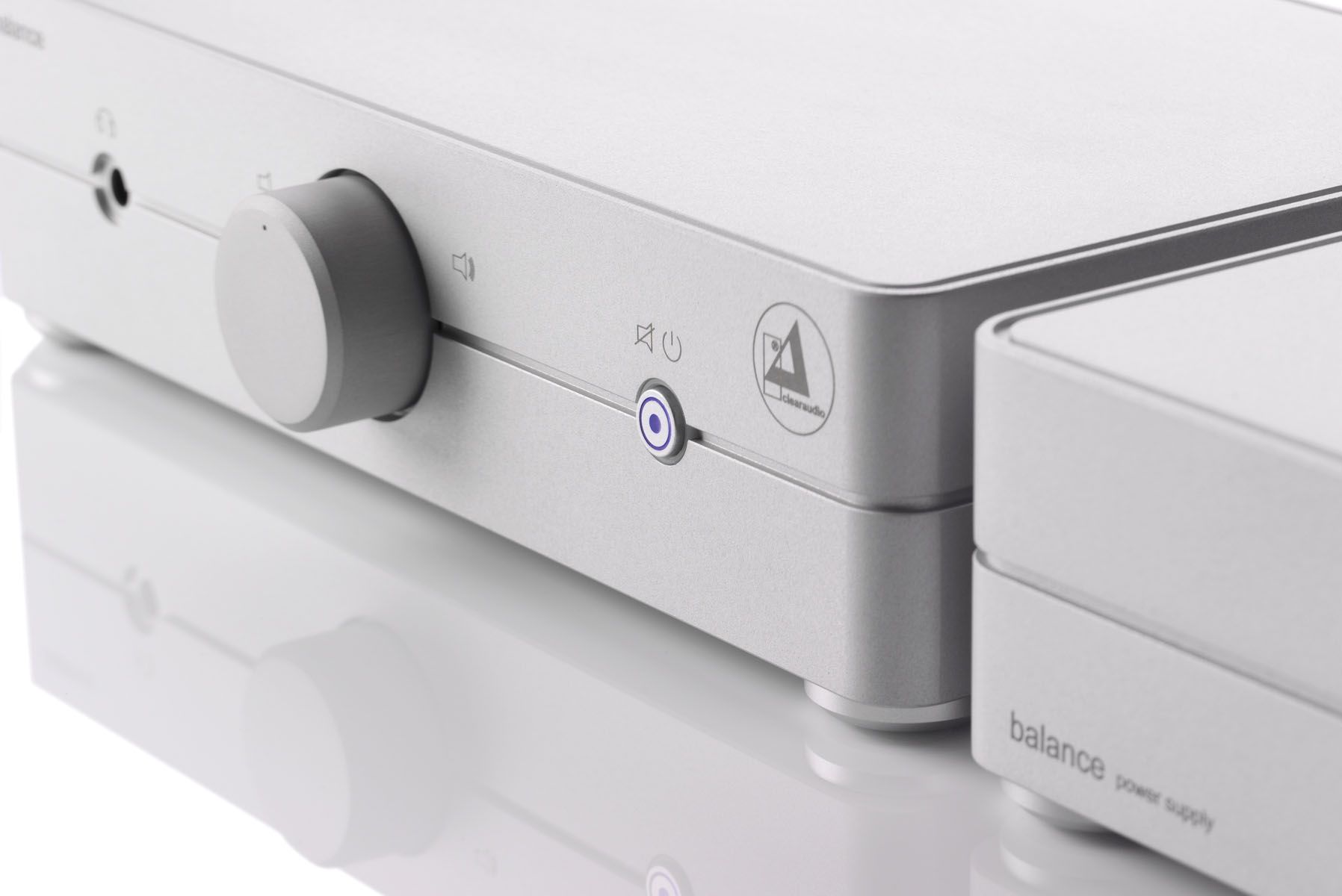 Perspektivische Nahaufnahme der silbernen Frontpartie der clearaudio balance V2 Phonovorstufe mit zentralem Drehregler, blau leuchtendem Ring-Einschaltknopf und dem danebenstehenden externen Netzteil „balance power supply“.