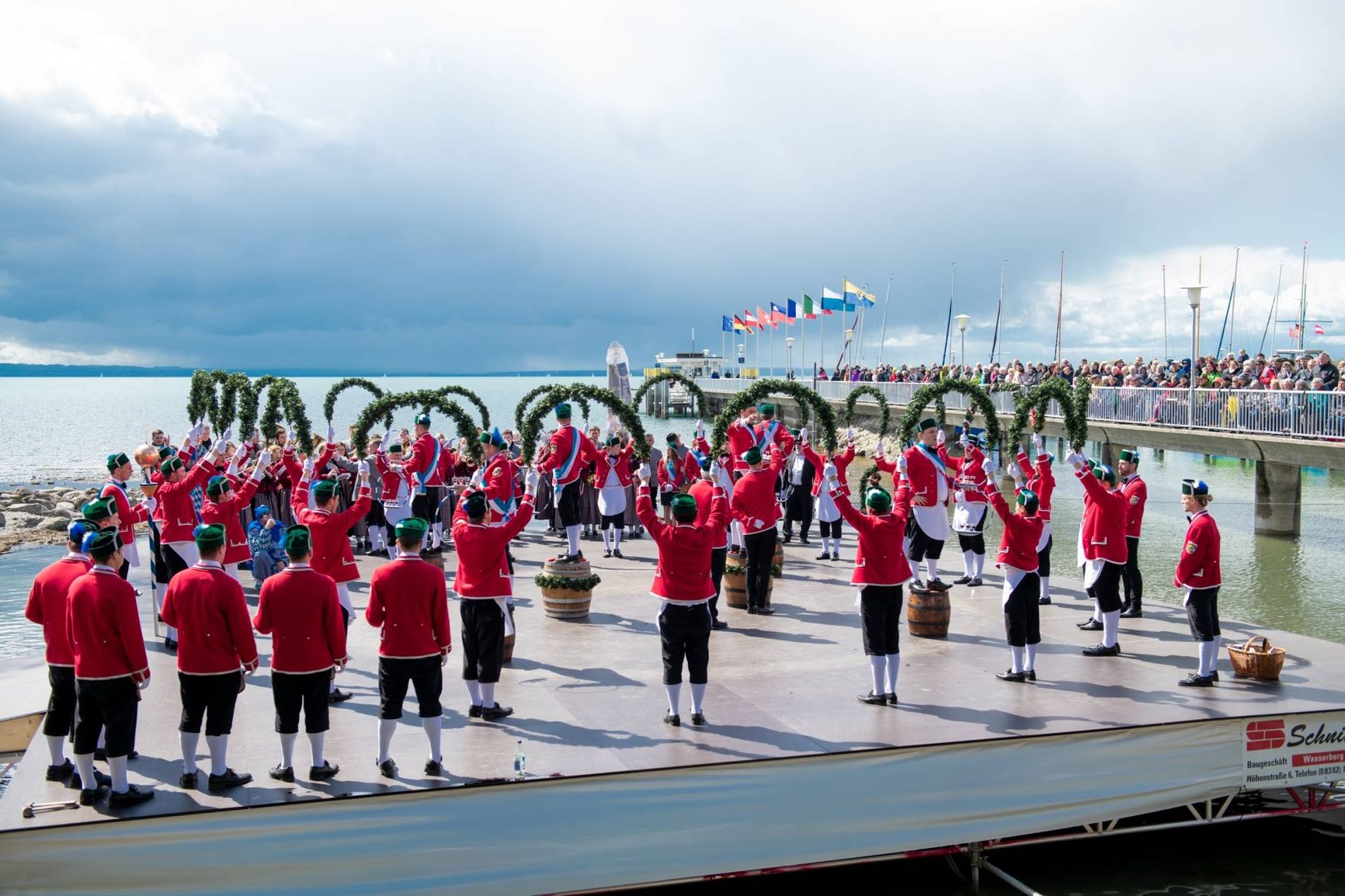 Man sieht die Bühne im Wasser und die Zuschauer auf dem Landesteg Die Schäffler bilden Formationen mit Kränzen, die Reifenschwinger stehen auf den Fässern.