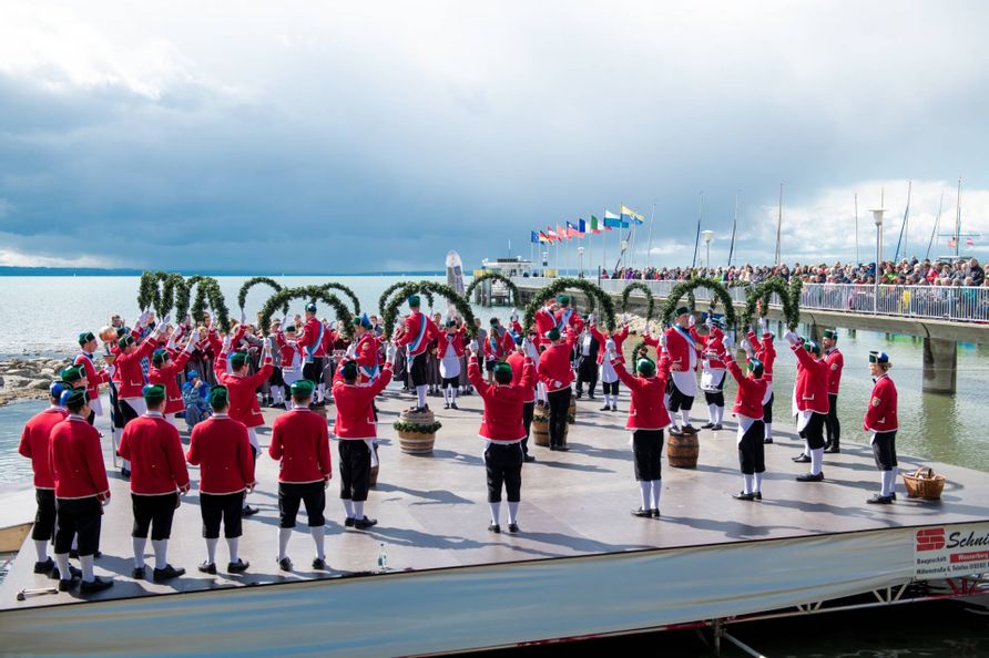 Man sieht die Bühne im Wasser und die Zuschauer auf dem Landesteg Die Schäffler bilden Formationen mit Kränzen, die Reifenschwinger stehen auf den Fässern.