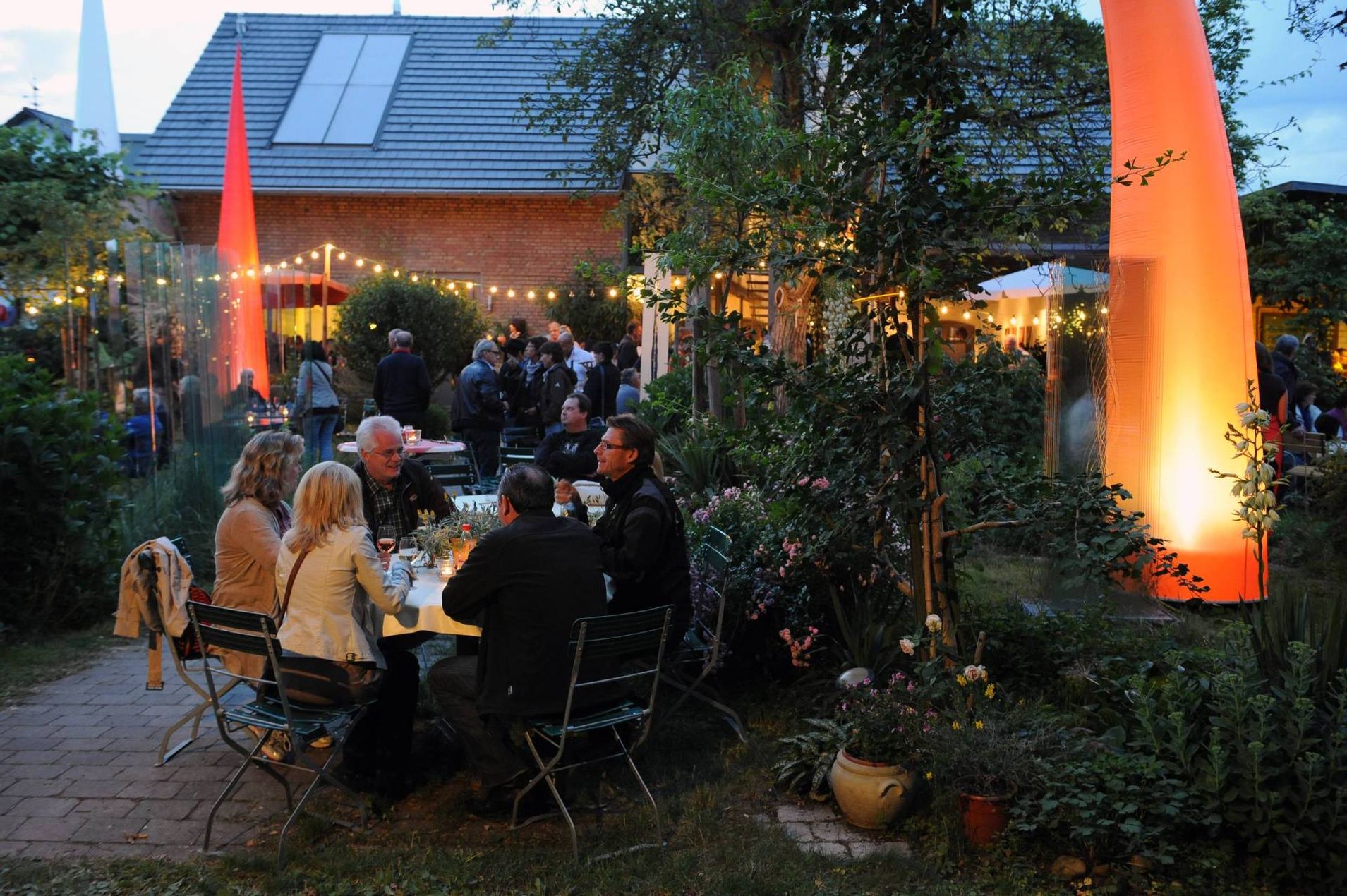 Schön beleuchteter Garten mit Tischen und Stühlen und Feiernden an Komm und See