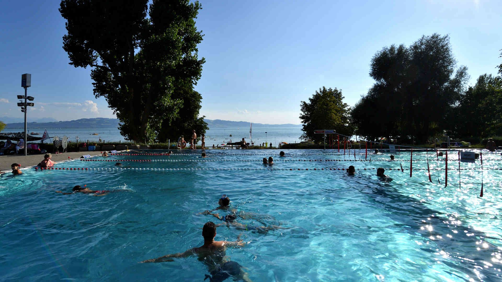 Liegewiese, Schwimmbecken und Seezugang im Freibad Aquamarin in Wasserburg. Im Hintergrund ist der See und die Bergwelt zu sehen.