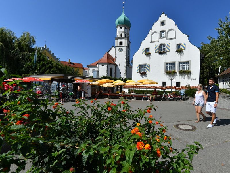 Ein Pärchen flaniert auf der Halbinsel in Wasserburg. Im Hintergrund ist die Kirch St. Georg, der Kiosk und das Museum im Malhaus zu sehen.