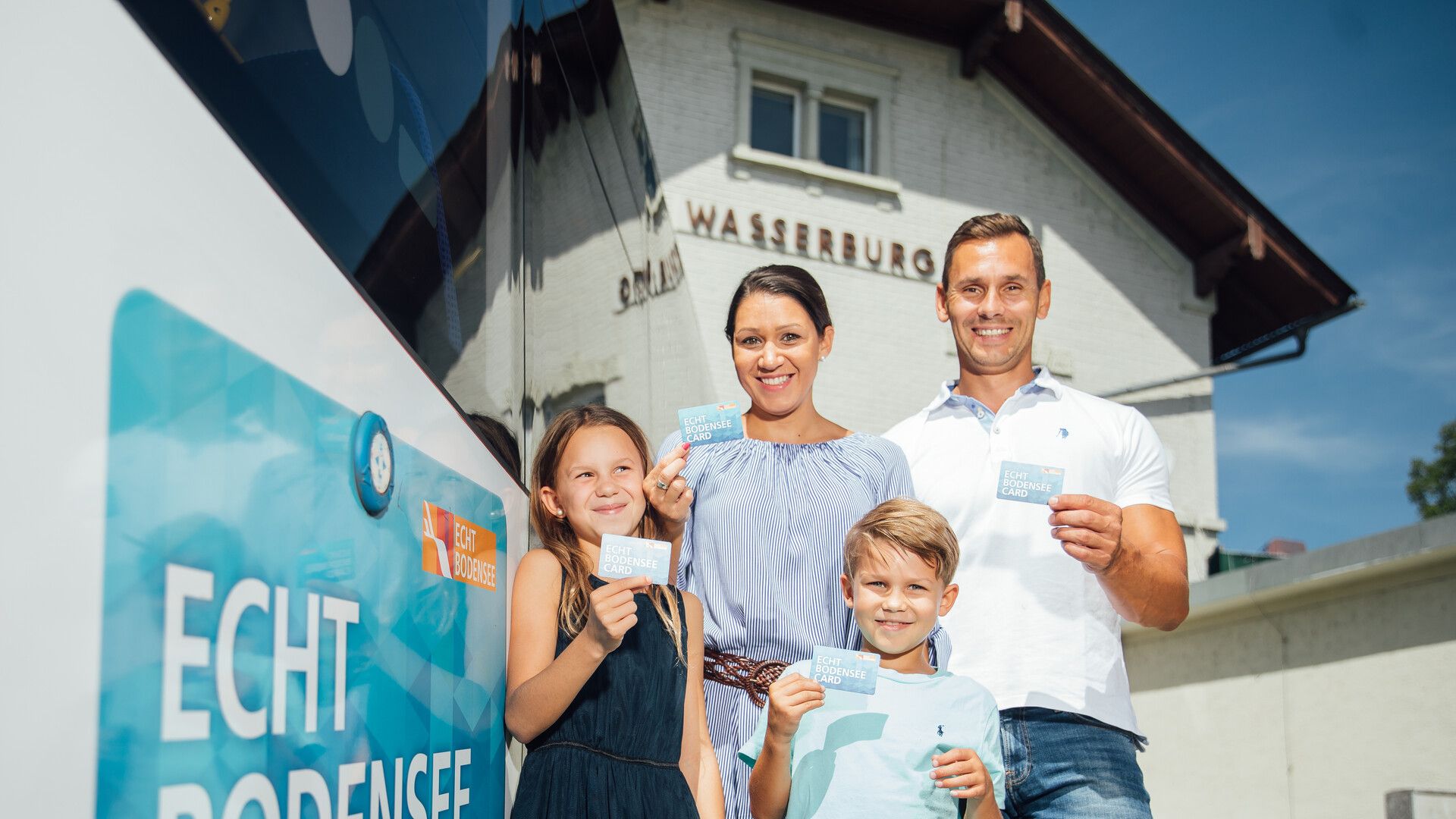 Eine Familie mit zwei Kindern vor dem Bahnhof in Wasserburg. Sie halten ihre Echt Bodensee Card in der Hand.