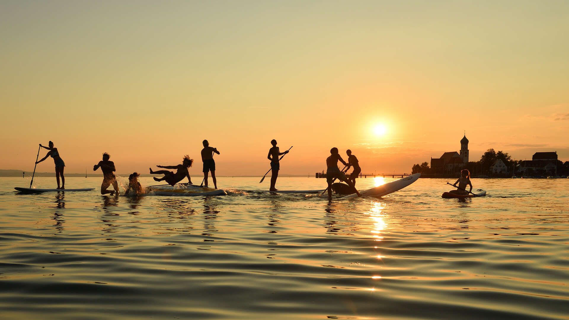 Mehrere Jugendliche mit ihrem SUP auf dem Bodensee vor der Halbinsel in Wasserburg. Im Hintergrund ist der Sonnenuntergang zu sehen.