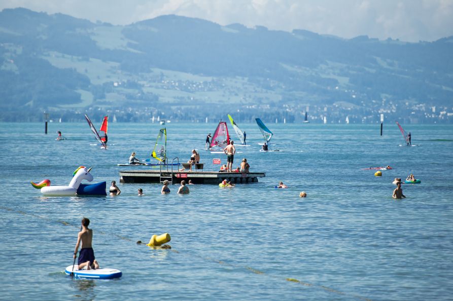 Floss Vom Freibad Aquamarin im Bodensee. Drum herum ein paar Windsurfer. Im Hintergrund ist die Bergwelt zu sehen.