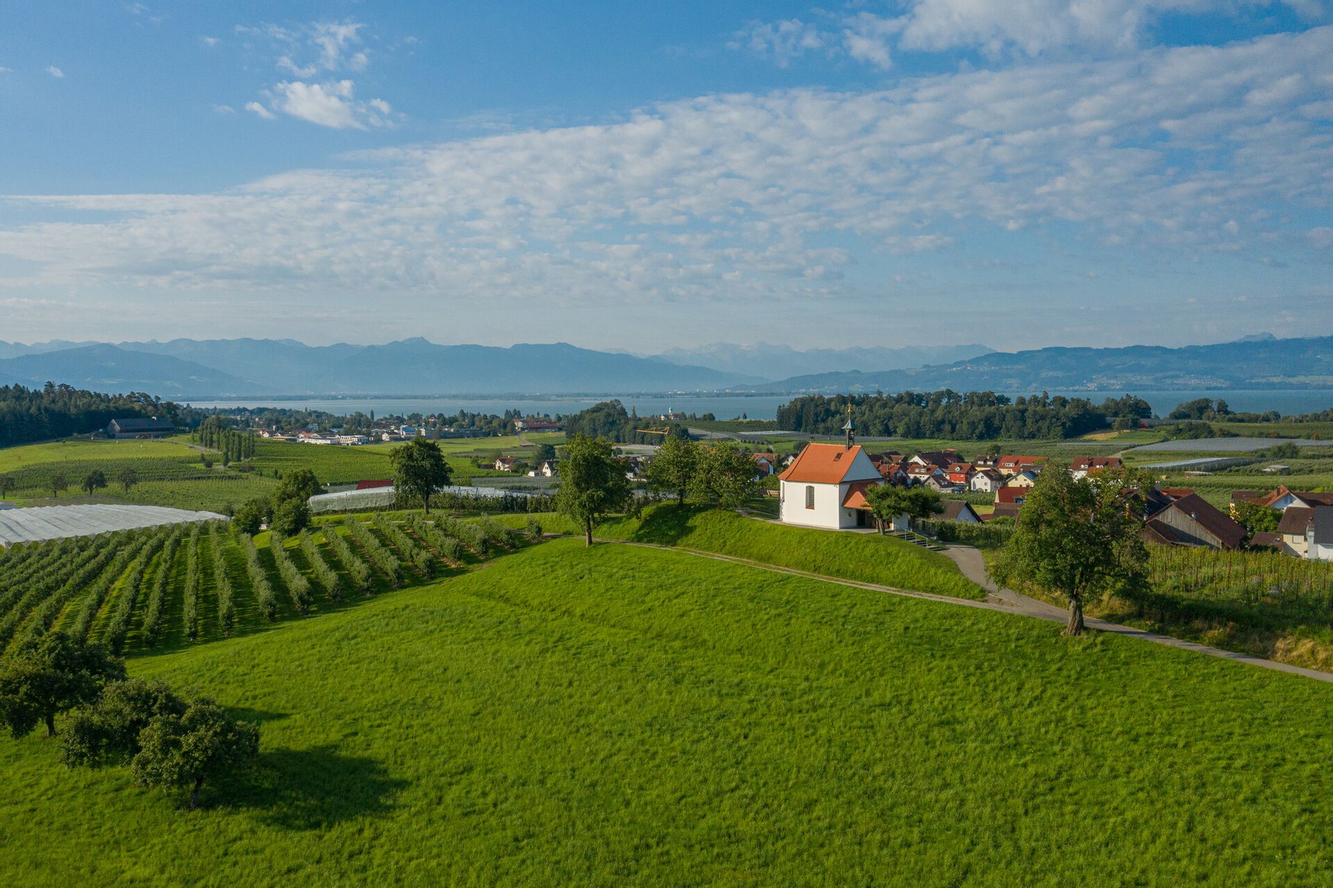 Luftbild Antoniuskapelle umringt von Feldern. Im Hintergrund ist der See und die Bergwelt zu sehen.