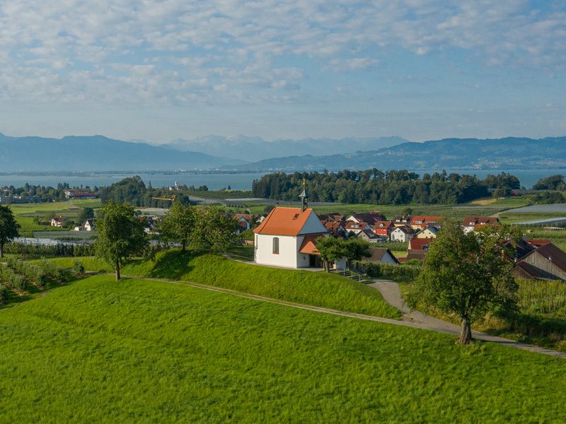 Luftbild Antoniuskapelle umringt von Feldern. Im Hintergrund ist der See und die Bergwelt zu sehen.