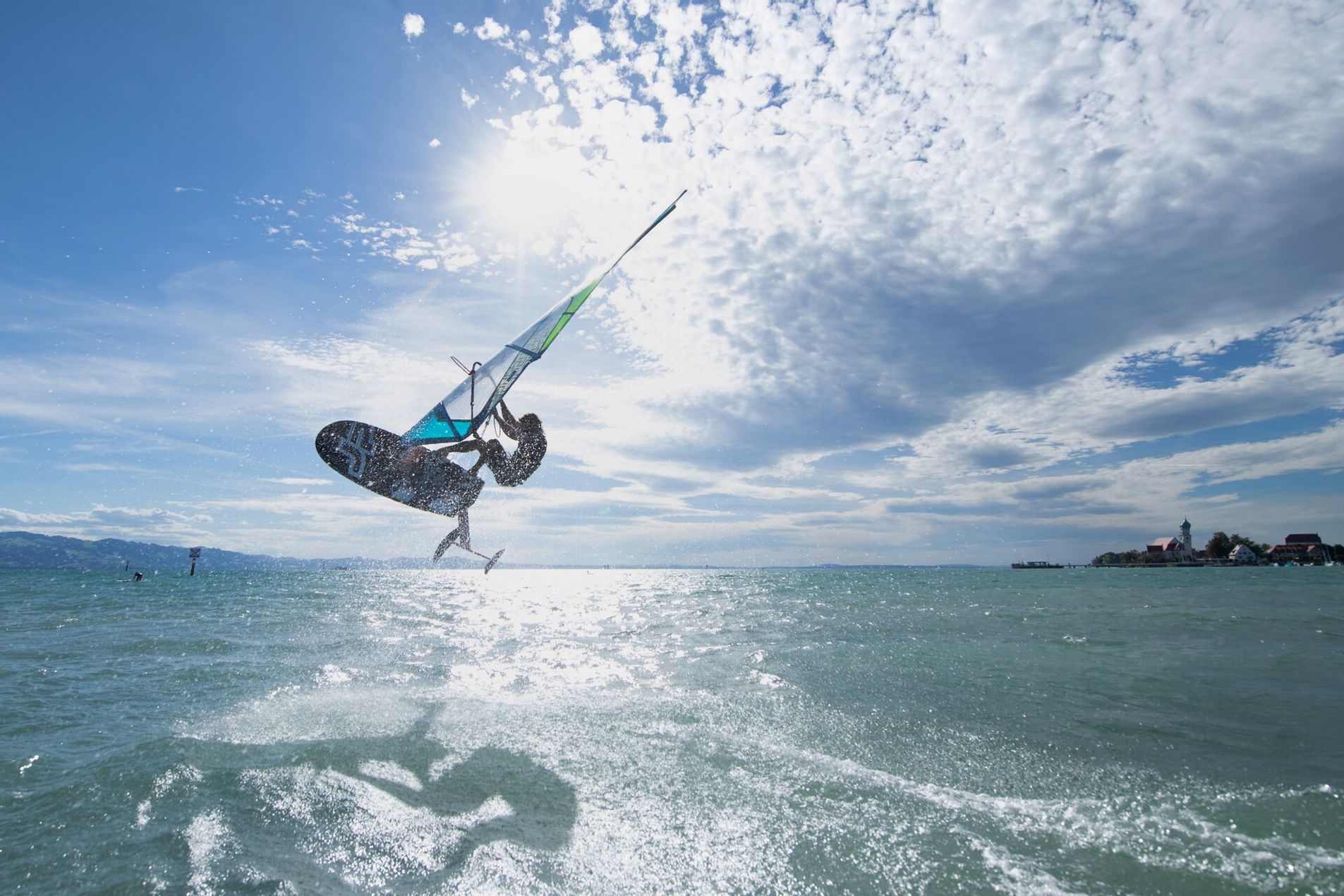 Ein Windsurfer auf dem Bodensee jumpt in höhe. Im Hintergrund ist die Halbinsel in Wasserburg zu sehen.