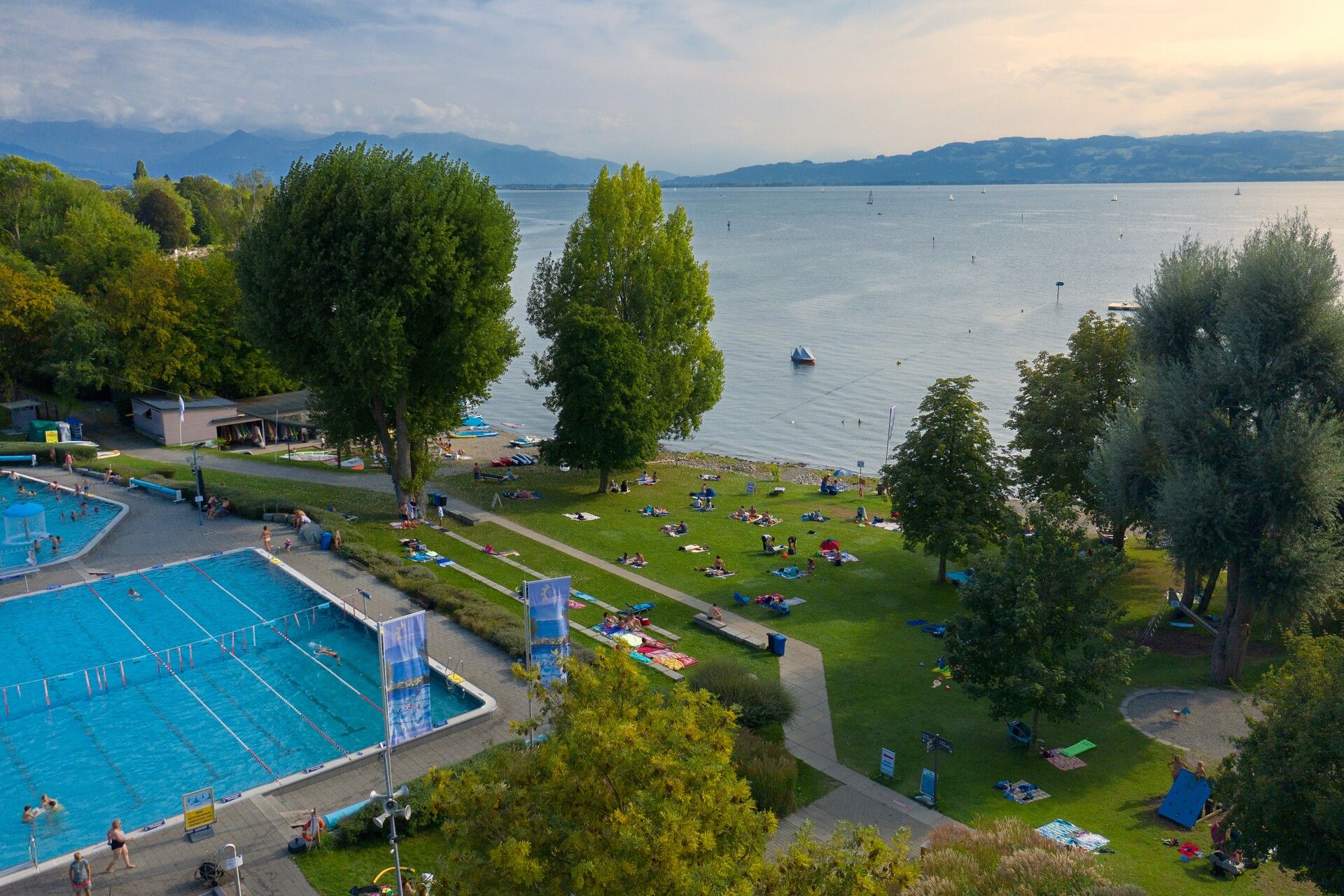 Luftaufnahme Freibad Aquamarin mit Beckenbereich, Liegewiese und dem Seezugang. Im Hintergrund ist die Bergwelt zu sehen.