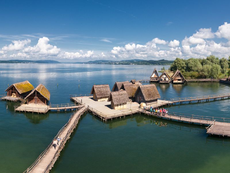Luftbild Pfahlbaumuseum Unteruldingen umringt vom Bodensee. Im Hintergrund die Bergwelt.