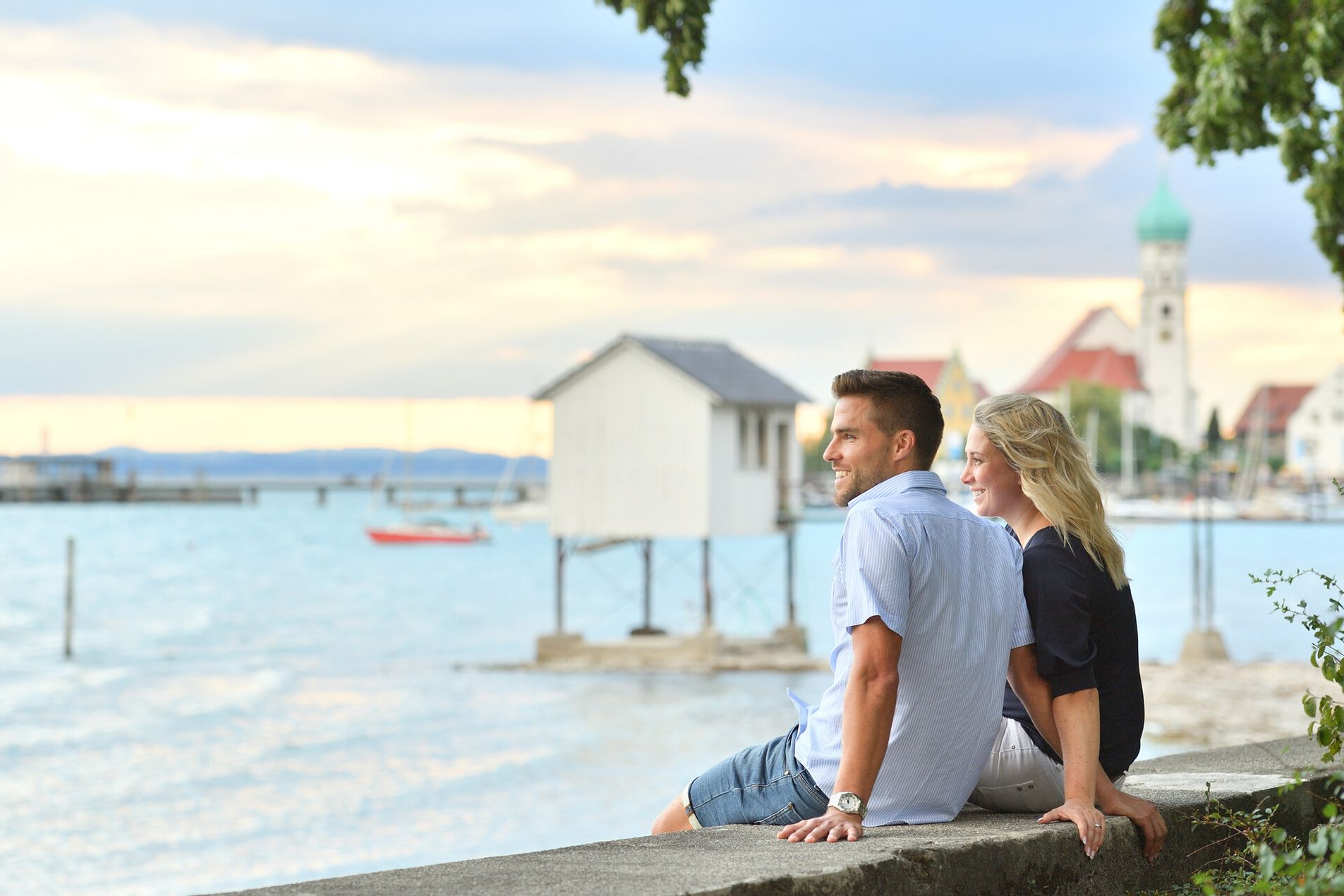 Eine Frau und ein Mann sitzen auf einer Mauer direkt am Bodensee in Wasserburg. Im Hintergrund ist verschwommen die Halbinsel zu sehen.