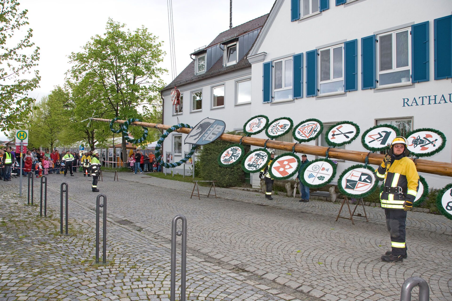 Maibaumstellen, der Maibaum liegt vor dem Rathaus. Feuerwehrleute stehen drum herum um ihn hochzuziehen.