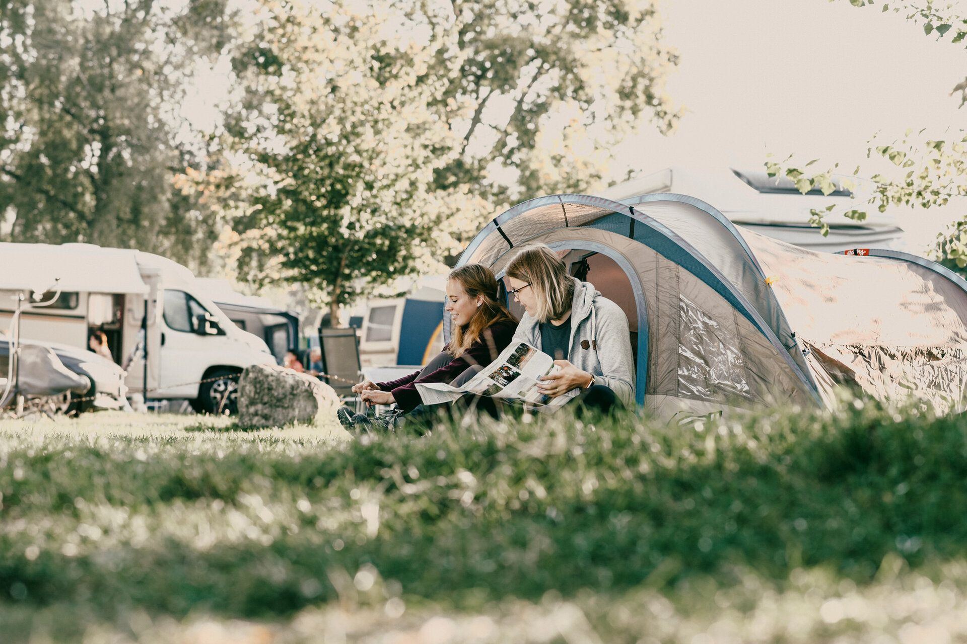 Zwei Frauen sitzen vor einem Zelteingang und schauen sich Prospekte vom Bodensee an. Im Hintergrund ist der Campingplatz zu sehen.