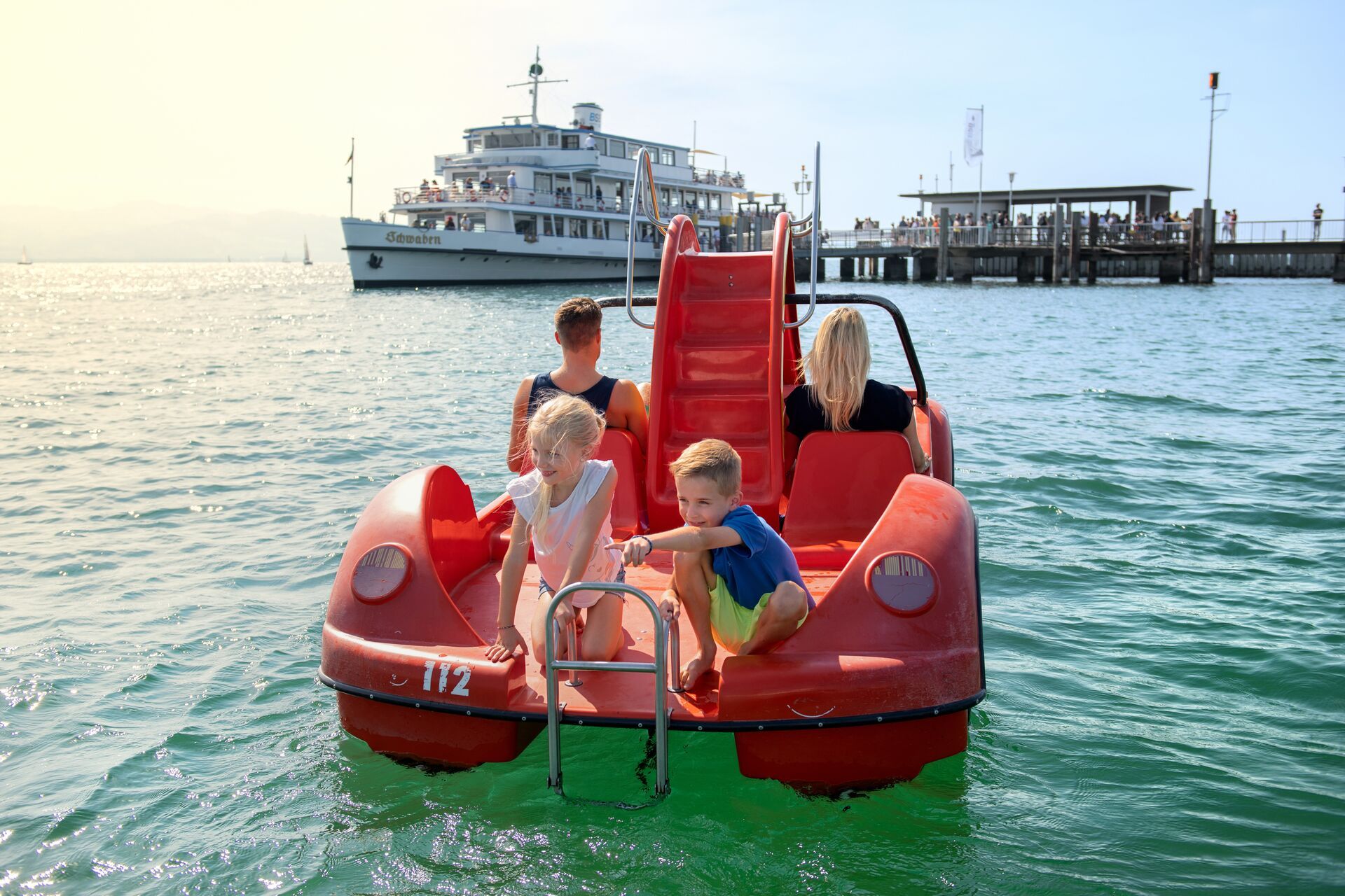 Eine Familie mit zwei Kindern auf einem Tretboot vor der Hafeneinfahrt in Wasserburg. Im Hintergrund ein Schiff der Weißenflotte vom Bodensee.