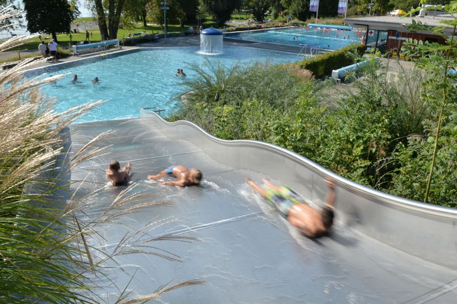 Kinder die die Wasserrutsche runter Rutschen im Freibad Aquamarin in Wasserburg. Im Hintergrund ist das Schwimmbecken und die Liegewiese zu sehen.