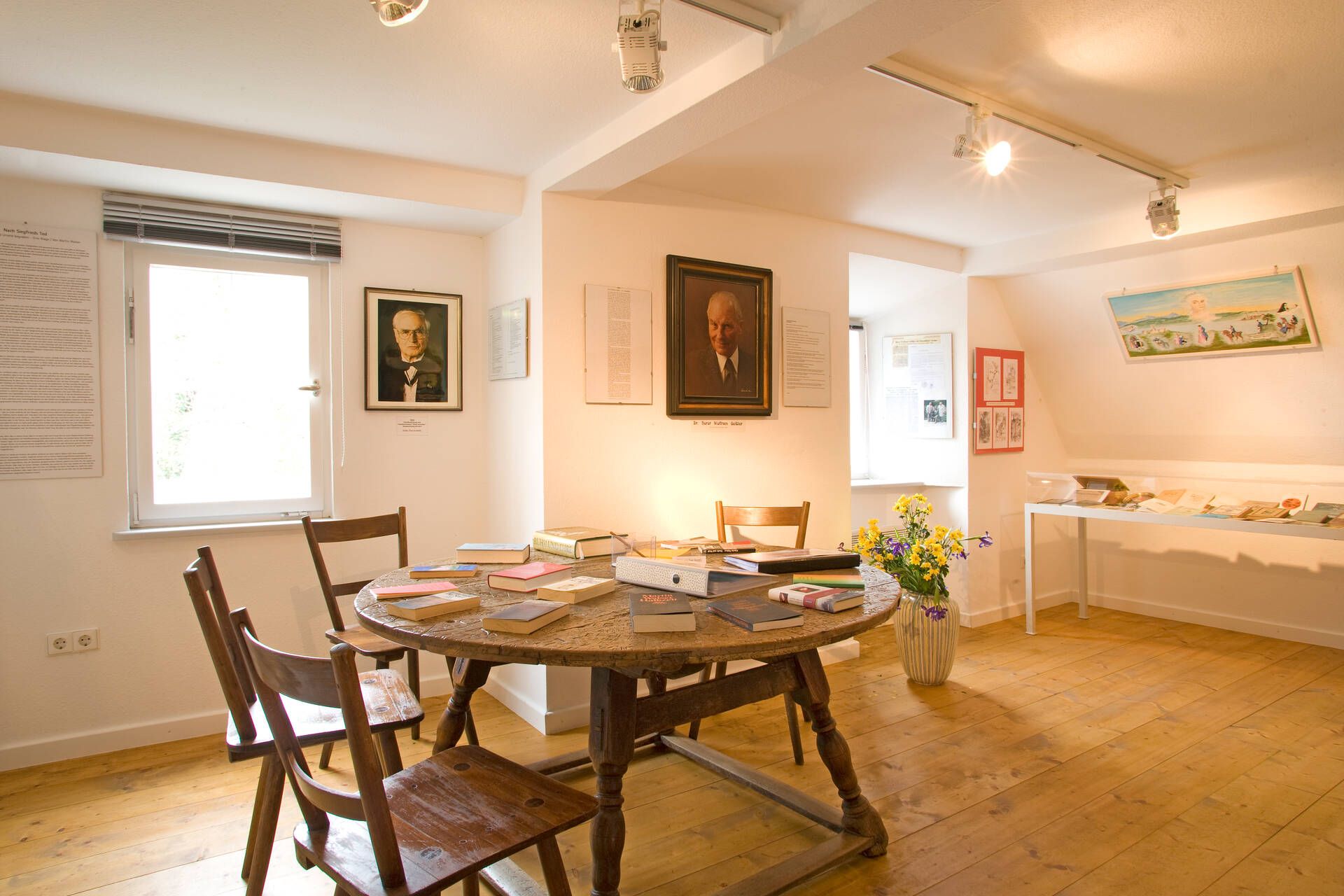 Ein Raum im Museum Malhaus in Wasserburg. Es ist ein Runder Tisch mit Büchern darauf zu sehen. An der Wand befinden sich Gemälde.