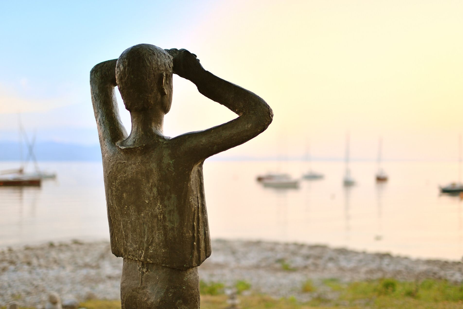 Das Denkmal des lieben Augustin in Wasserburg. Im Hintergrund ist verschwommen der Bodensee mit Segelboten zu sehen.